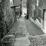 1960 :  M. Collinet bavarde avec une petite fille dans une rue qui a subi beaucoup de travaux : des pavés ont été remplacés par de l’asphalte, l’avaloir n’a pas été reconstitué et les pavés anciens sont mal assemblés.